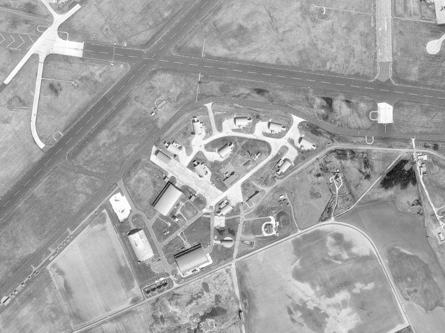 A historic black and white vertical aerial photo of an airfield. 2 runways cross in an X shape at the tope of the image. In the middle of the image are several concrete bunkers grouped in a rough circle.