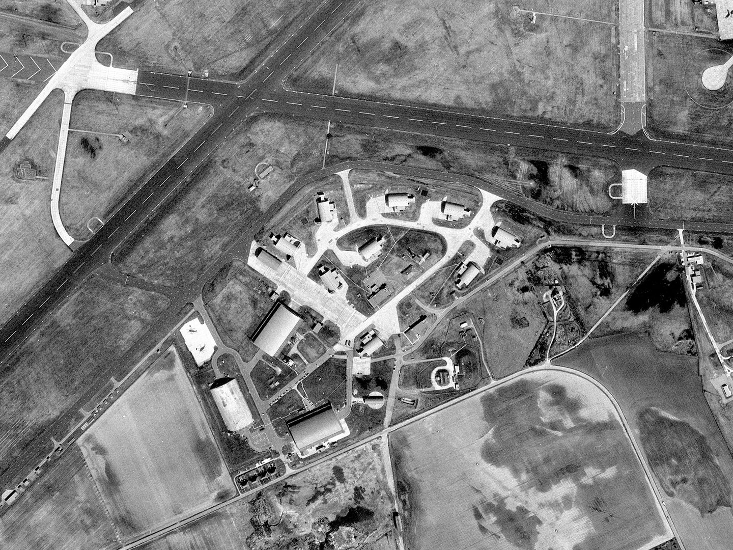 A historic black and white vertical aerial photo of an airfield. 2 runways cross in an X shape at the tope of the image. In the middle of the image are several concrete bunkers grouped in a rough circle.