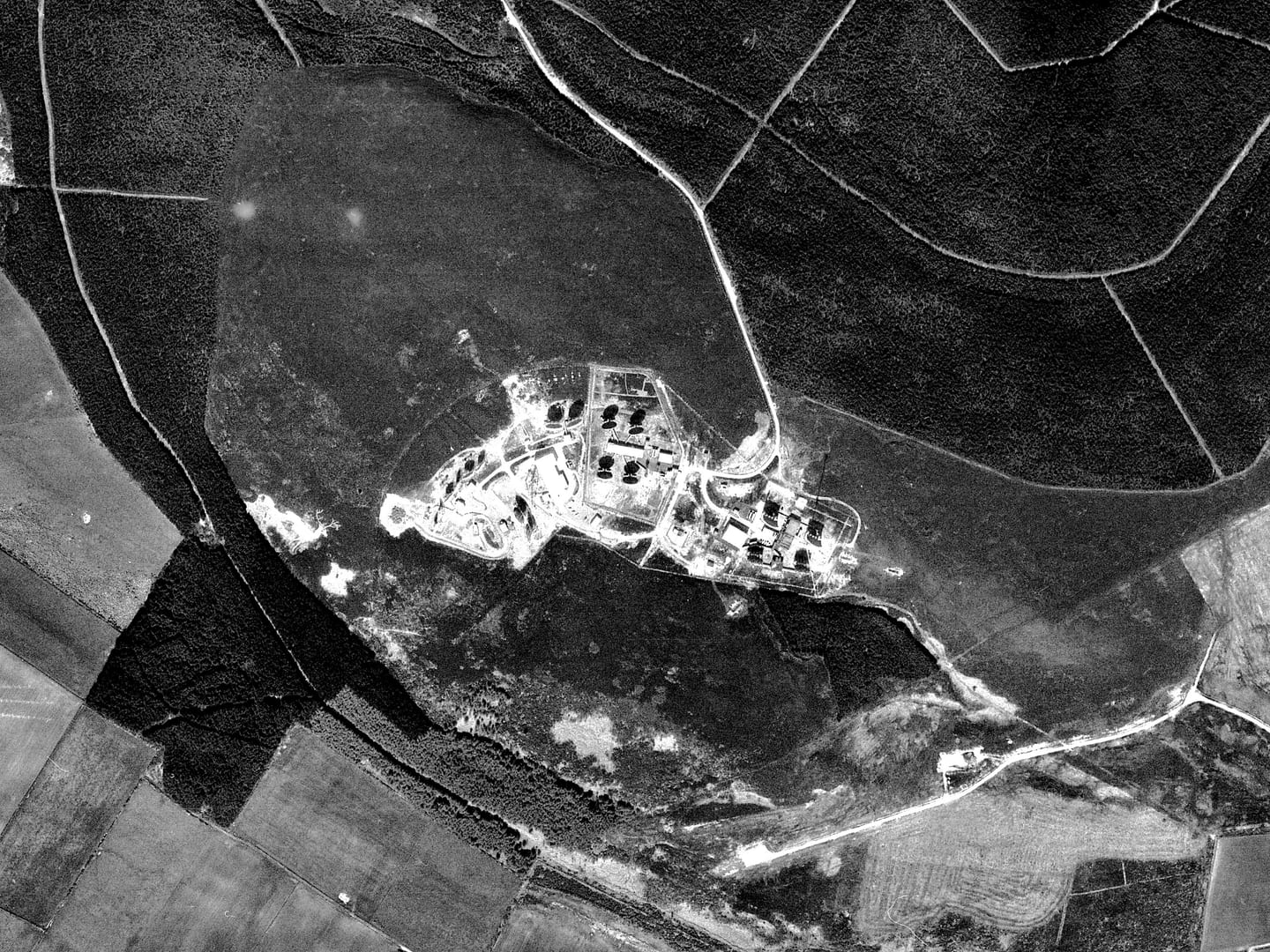 A black and white photo aerial photo of a wood with a network of white tracks running through it. There is a clearing in the middle containing a complex of buildings and structures. Some of these are casting stark black shadows.