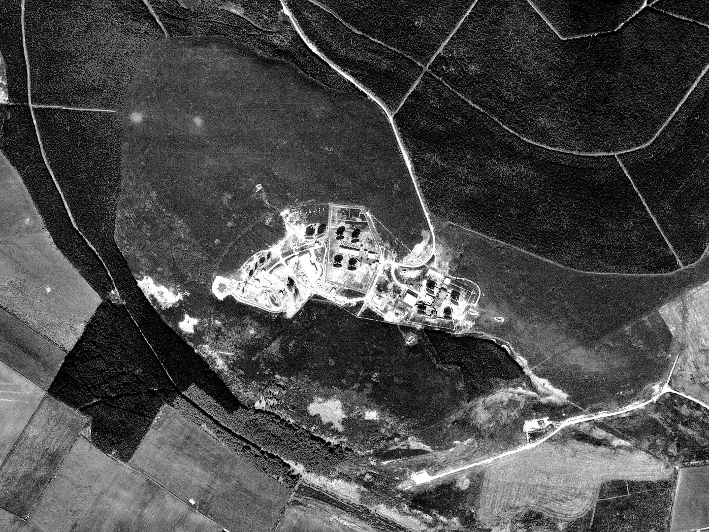 A black and white photo aerial photo of a wood with a network of white tracks running through it. There is a clearing in the middle containing a complex of buildings and structures. Some of these are casting stark black shadows.