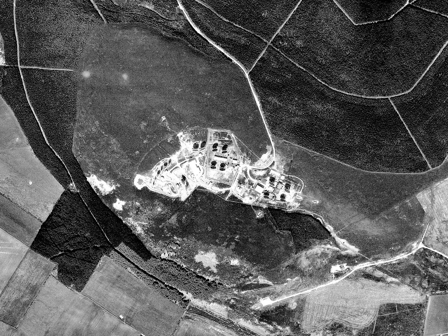 A black and white photo aerial photo of a wood with a network of white tracks running through it. There is a clearing in the middle containing a complex of buildings and structures. Some of these are casting stark black shadows.