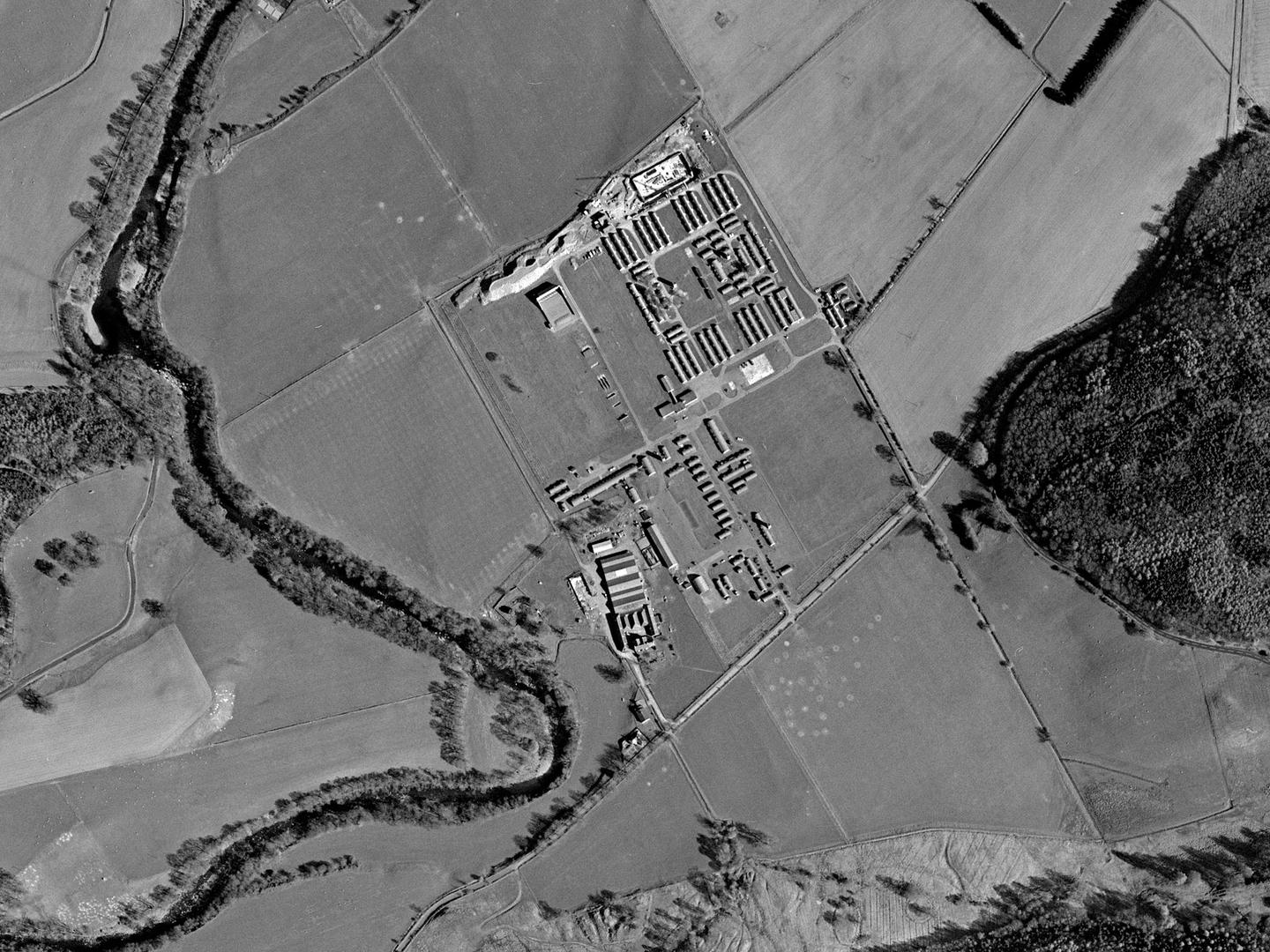 A black and white aerial photo of a landscape with a river meandering vertically through the left half. The landscape is comprised of fields and small woodlands. A camp with rows of huts is in the middle, with excavations just visible to its north.