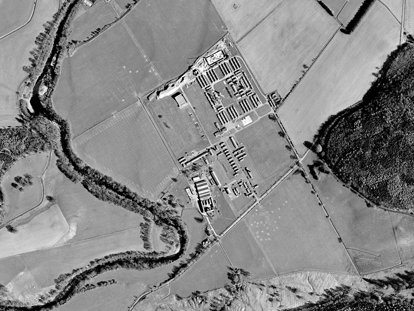 A black and white aerial photo of a landscape with a river meandering vertically through the left half. The landscape is comprised of fields and small woodlands. A camp with rows of huts is in the middle, with excavations just visible to its north.