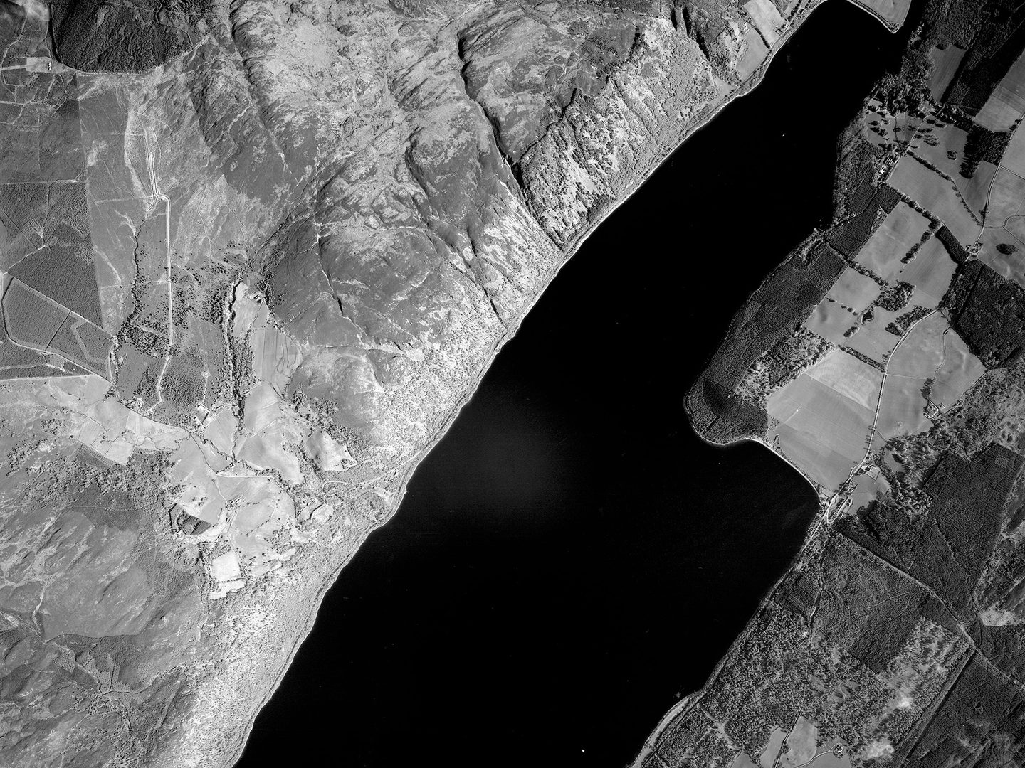 A black and white vertical aerial photo of Loch Ness. The black waters of the loch run diagonally from the top right corner to the bottom of the image. North of the loch is a steep, wooded shore and rocky moorland. The southern shore is heavily wooded.