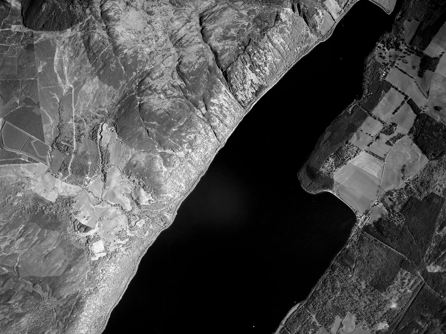 A black and white vertical aerial photo of Loch Ness. The black waters of the loch run diagonally from the top right corner to the bottom of the image. North of the loch is a steep, wooded shore and rocky moorland. The southern shore is heavily wooded.