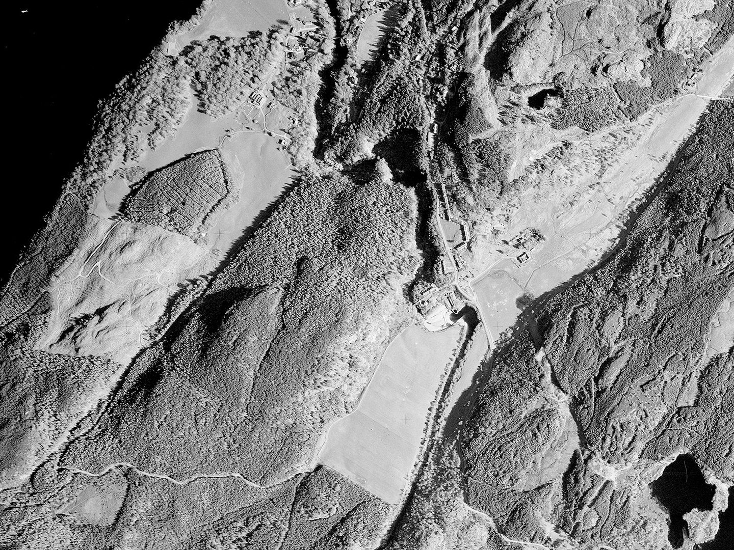 A historic black and white vertical aerial photo of a wooded landscape. A small dam and series of buildings are visible in the centre of the image. A small body of water is visible in the bottom right corner, with a larger body of water top left.