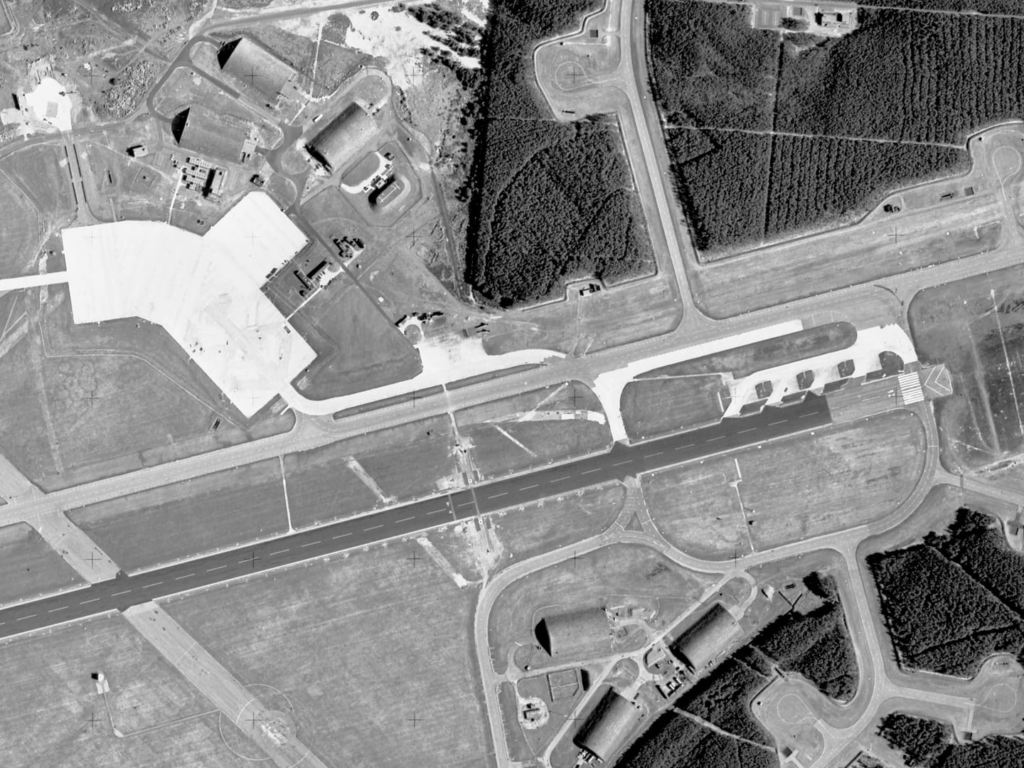 A black and white aerial photo of an airfield. A runway runs horizontally through the middle of the image. At the right-hand end are 4 concrete stands. The runway is flanked by trees and 6 large hangars – 3 above and 3 below.