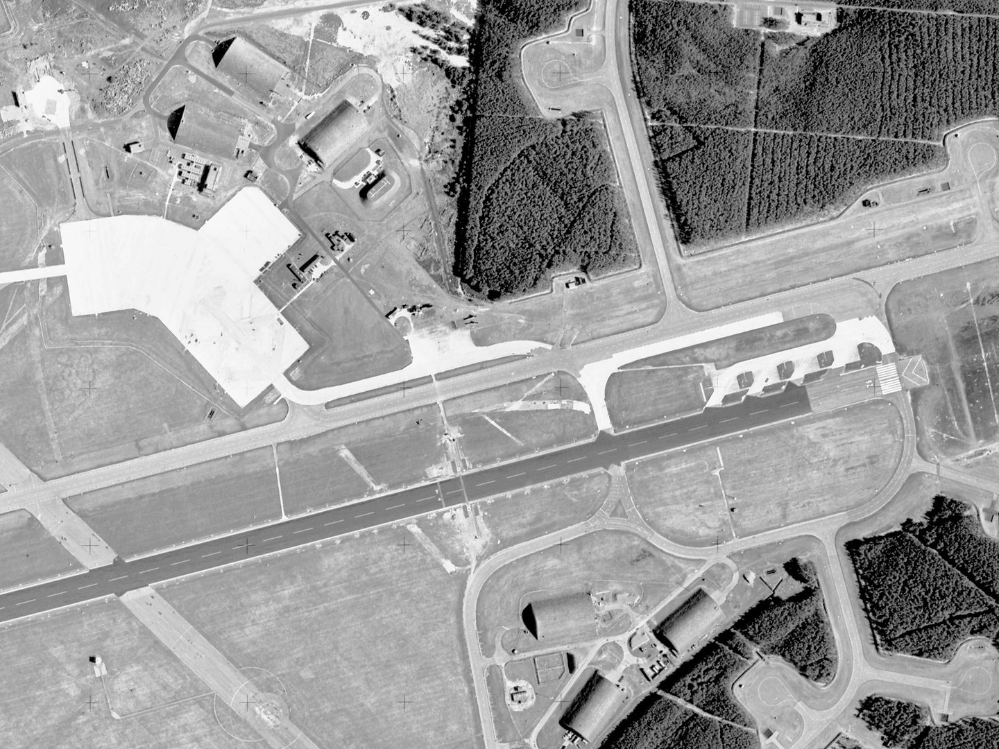 A black and white aerial photo of an airfield. A runway runs horizontally through the middle of the image. At the right-hand end are 4 concrete stands. The runway is flanked by trees and 6 large hangars – 3 above and 3 below.