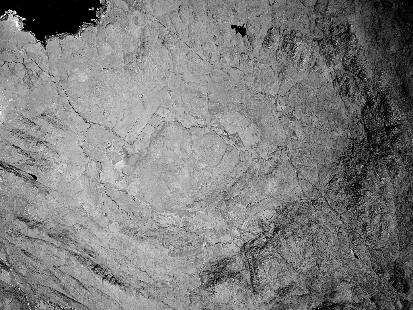 A black and white vertical aerial photo of empty moorland. It is surrounded by a huge, roughly oval ring of crags and rocks. A small patch of black sea is visible in the top left corner of the image.