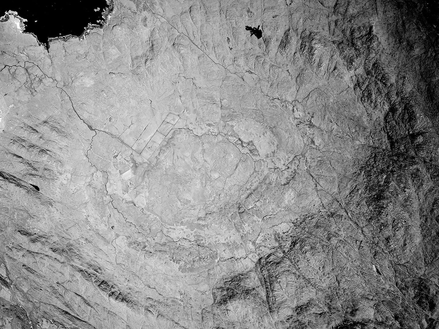 A black and white vertical aerial photo of empty moorland. It is surrounded by a huge, roughly oval ring of crags and rocks. A small patch of black sea is visible in the top left corner of the image.