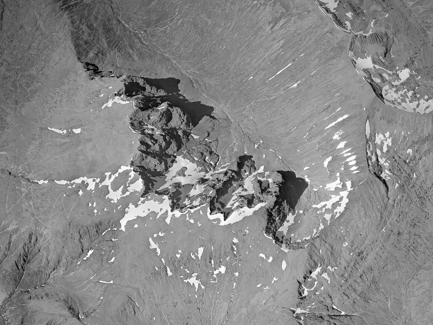 A black and white vertical aerial photo of the summit of Ben Nevis. The rocky peak is casting shadows towards the east/right of the image. A broad valley sweeps down from the peak to the north/top. Small areas of snow scatter the summit.