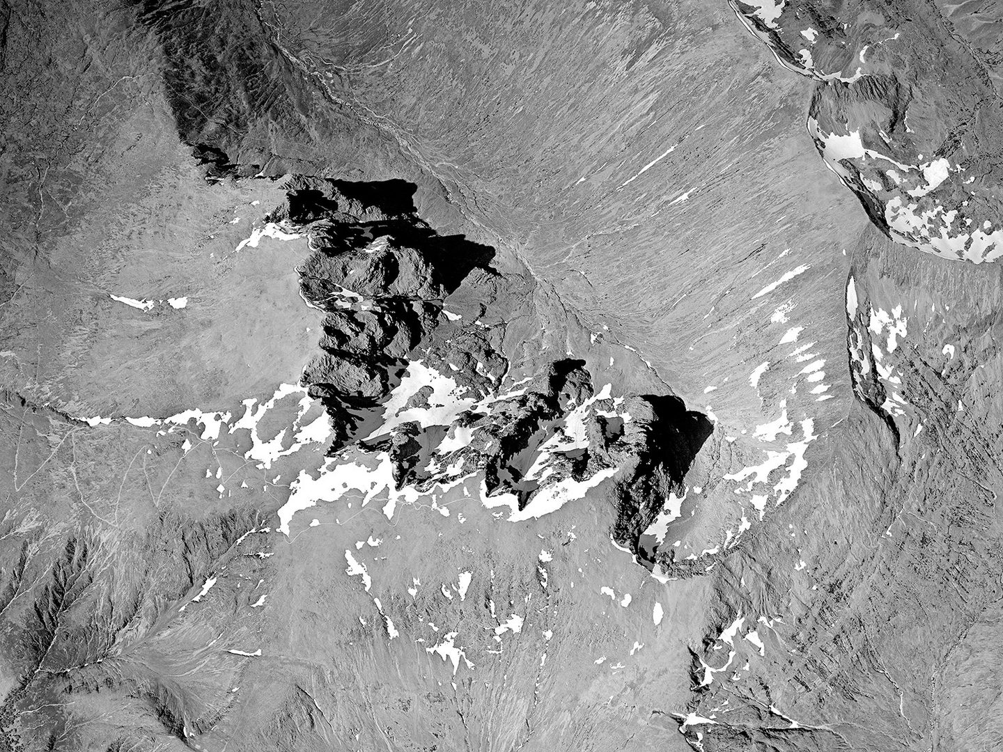 A black and white vertical aerial photo of the summit of Ben Nevis. The rocky peak is casting shadows towards the east/right of the image. A broad valley sweeps down from the peak to the north/top. Small areas of snow scatter the summit.