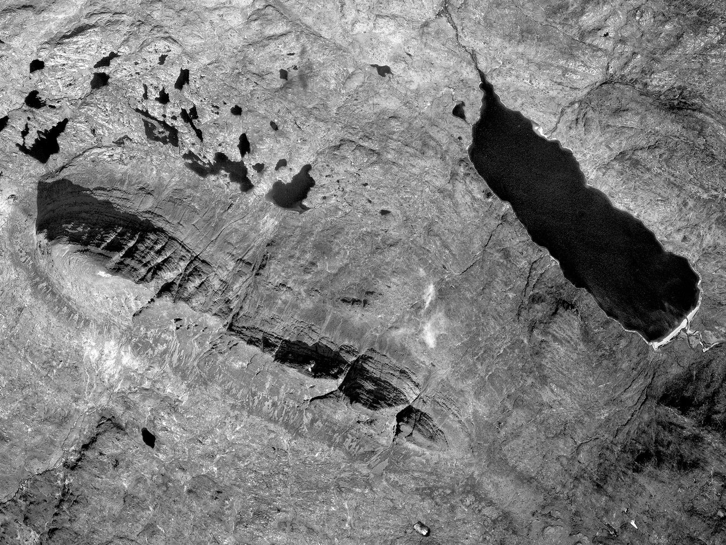 A black and white aerial photo of Suilven. The mountain forms a distinctive thin ridge, running diagonally across the image from the middle of the left edge to the middle of the bottom. A black loch runs parallel in the right part of the image.