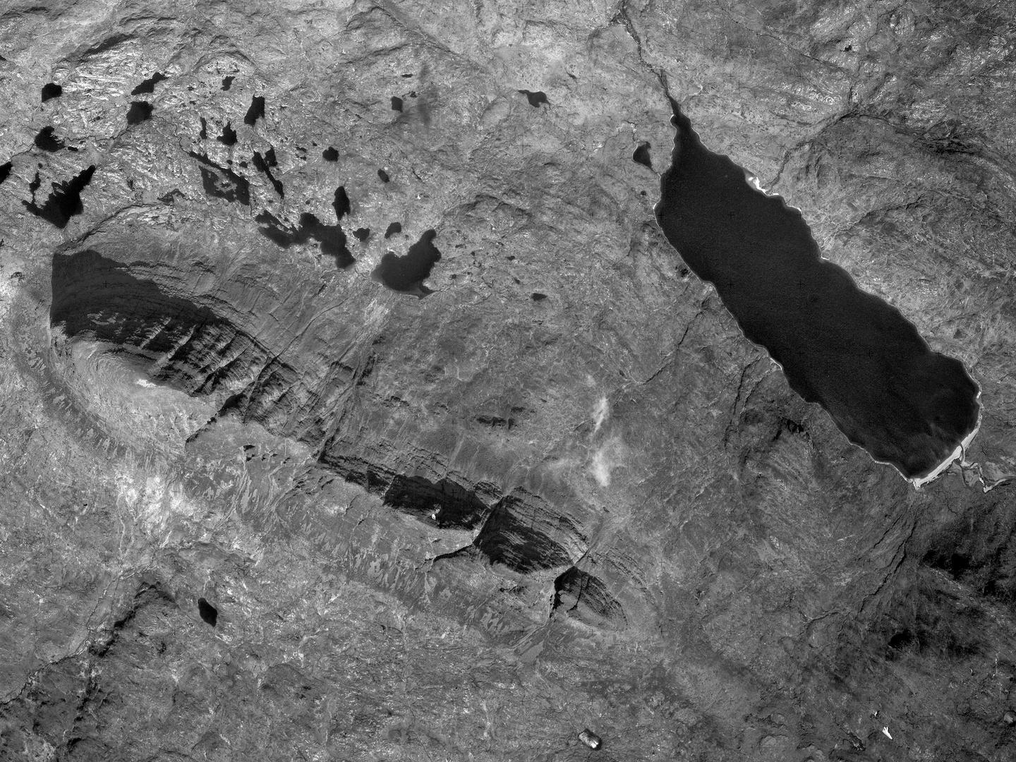 A black and white aerial photo of Suilven. The mountain forms a distinctive thin ridge, running diagonally across the image from the middle of the left edge to the middle of the bottom. A black loch runs parallel in the right part of the image.