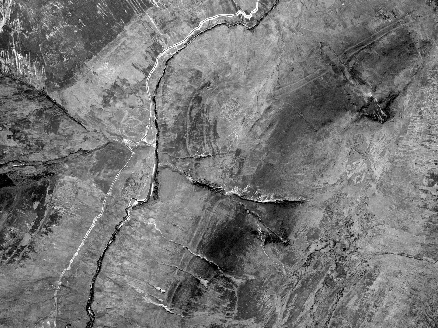 A black and white vertical aerial photo of a landscape in the Scottish Highlands. A river and a track run across the image. Parallel to them and to their right, 3 parallel lines snake clearly across the landscape.