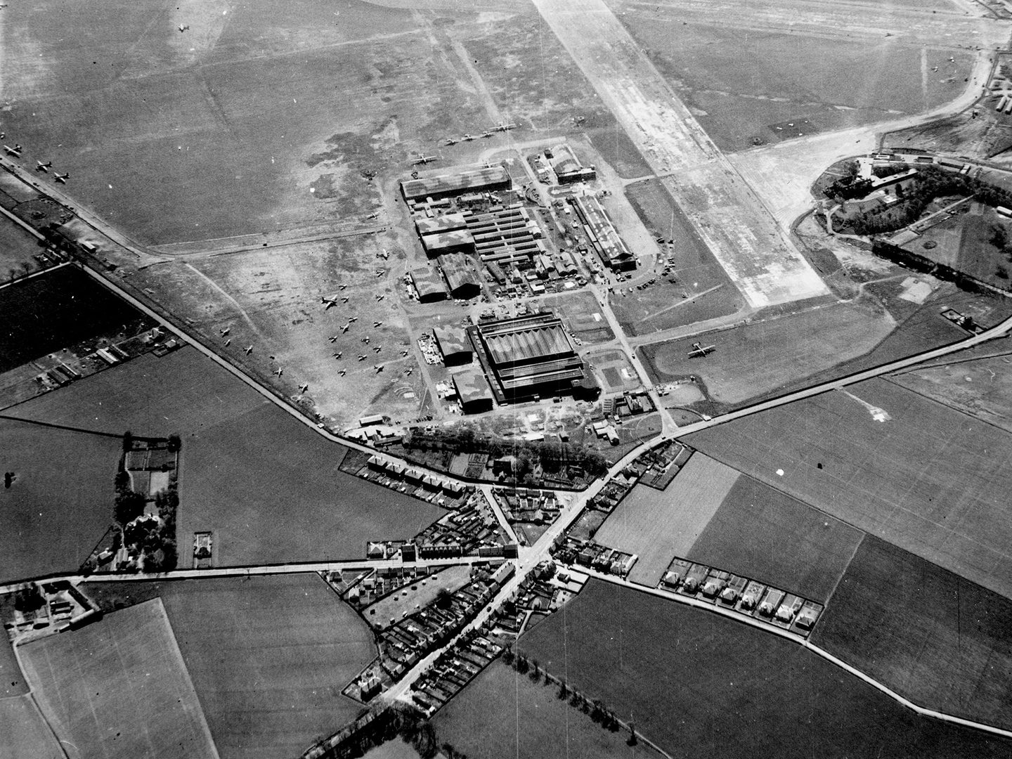 A historic black and white oblique aerial photo of an airfield. A large complex of buildings occupy the centre of the image, with several roads leading up to them. The buildings are surrounded by aircraft. A runway stretches out of the top edge of the frame.