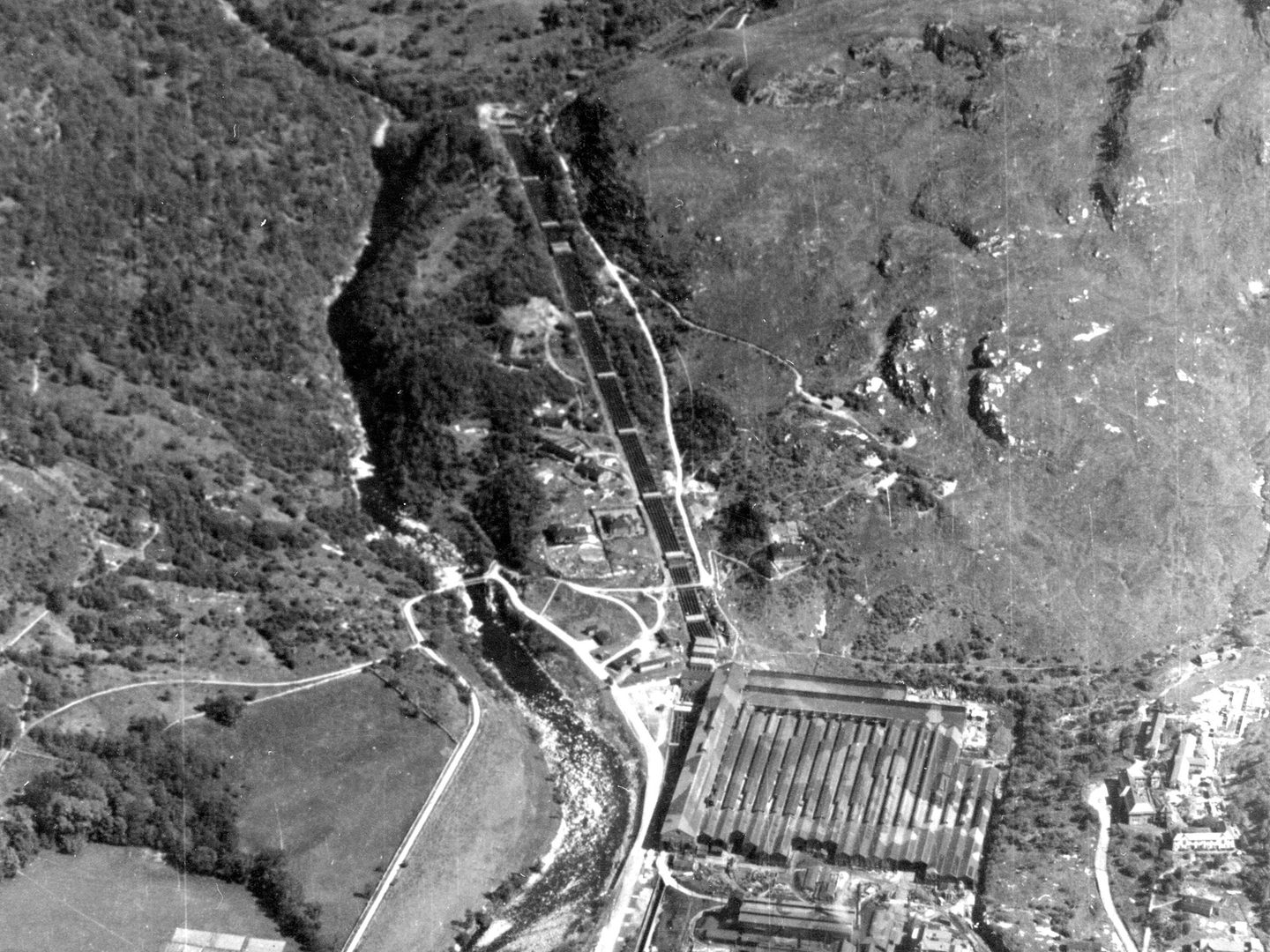 A historic black and white oblique aerial photo of a wooded hillside. A thick band of large pipes run downhill through the centre of the image to a large building at the bottom of the image. The roof of the building is painted in a camouflage pattern.