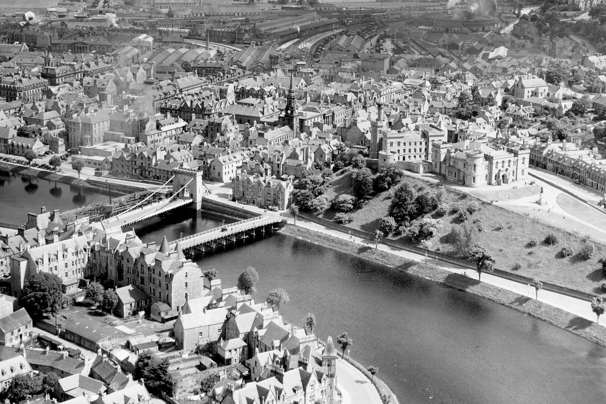 A historic black and white oblique aerial photo of Inverness. A river runs through the middle of the photo, with buildings on both banks. Two bridges cross the river. Inverness Castle is visible to the right of centre.