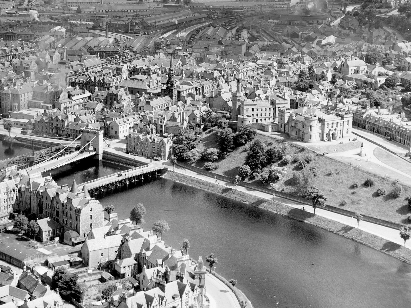 A historic black and white oblique aerial photo of Inverness. A river runs through the middle of the photo, with buildings on both banks. Two bridges cross the river. Inverness Castle is visible to the right of centre.