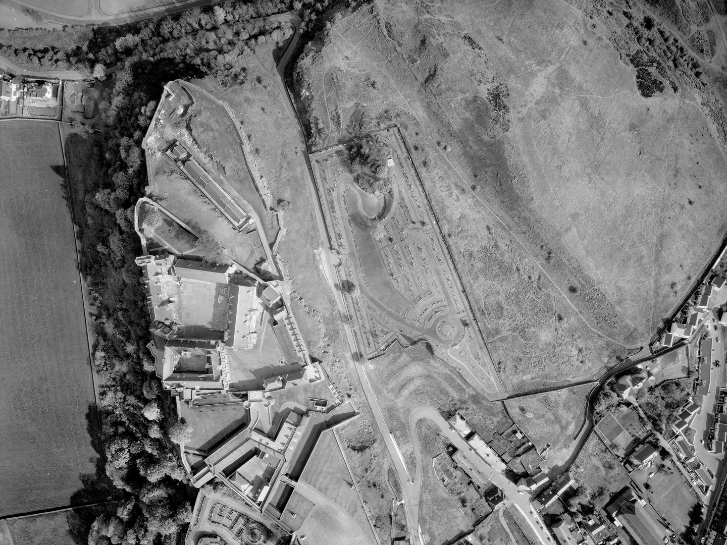 A black and white vertical aerial photo of Stirling Castle. The castle complex is on top of a steep crag, with wooded slopes falling away to the left. To the right is a cemetery and open grassland. In the bottom right corner are some houses.