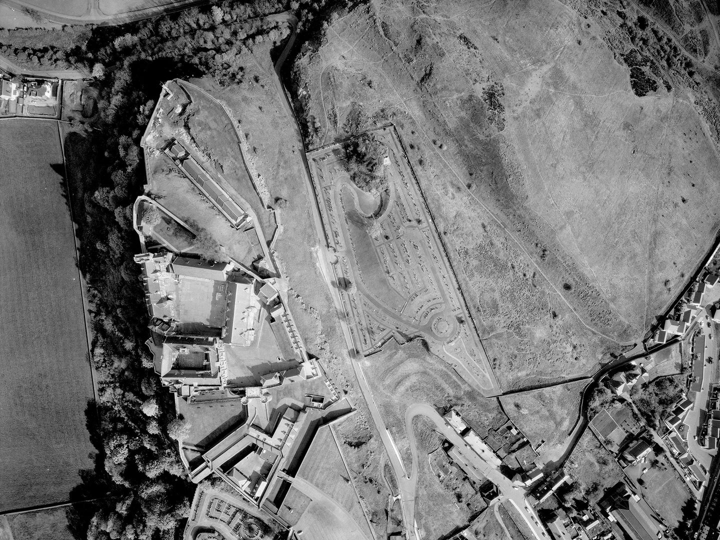 A black and white vertical aerial photo of Stirling Castle. The castle complex is on top of a steep crag, with wooded slopes falling away to the left. To the right is a cemetery and open grassland. In the bottom right corner are some houses.