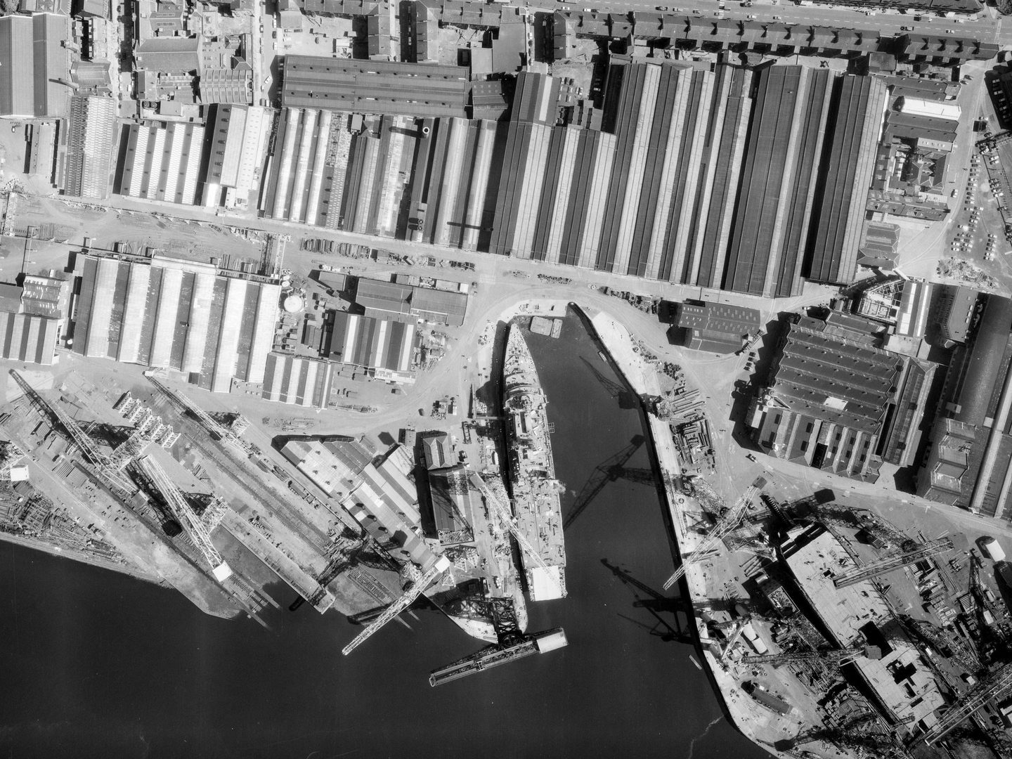 A black and white aerial photo of a port. A small amount of water is visible at the bottom edge, with a large ship tied up at a dock in the centre. There are cranes to the left of the ship and large warehouses across the dockside.