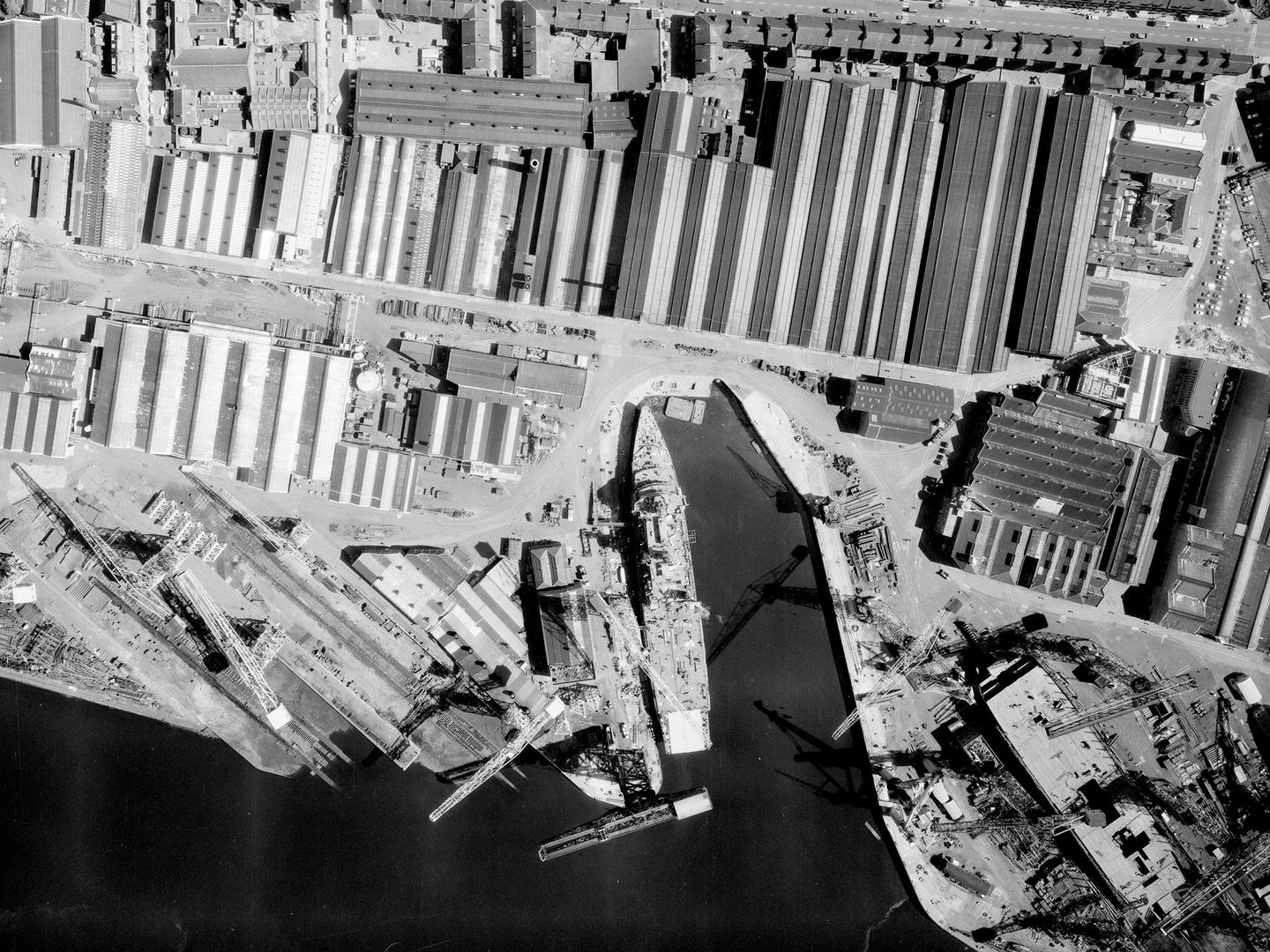 A black and white aerial photo of a port. A small amount of water is visible at the bottom edge, with a large ship tied up at a dock in the centre. There are cranes to the left of the ship and large warehouses across the dockside.