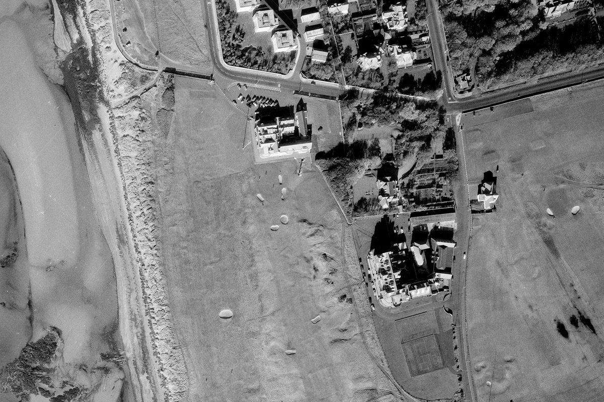 A historic black and white vertical aerial photo of a golf course along a shoreline. The water is to the left of the image, with the course running from top to bottom. Several grand buildings - including the Royal Troon clubhouse - are visible in the centre of the image.