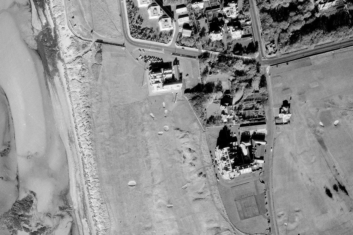 A historic black and white vertical aerial photo of a golf course along a shoreline. The water is to the left of the image, with the course running from top to bottom. Several grand buildings - including the Royal Troon clubhouse - are visible in the centre of the image.