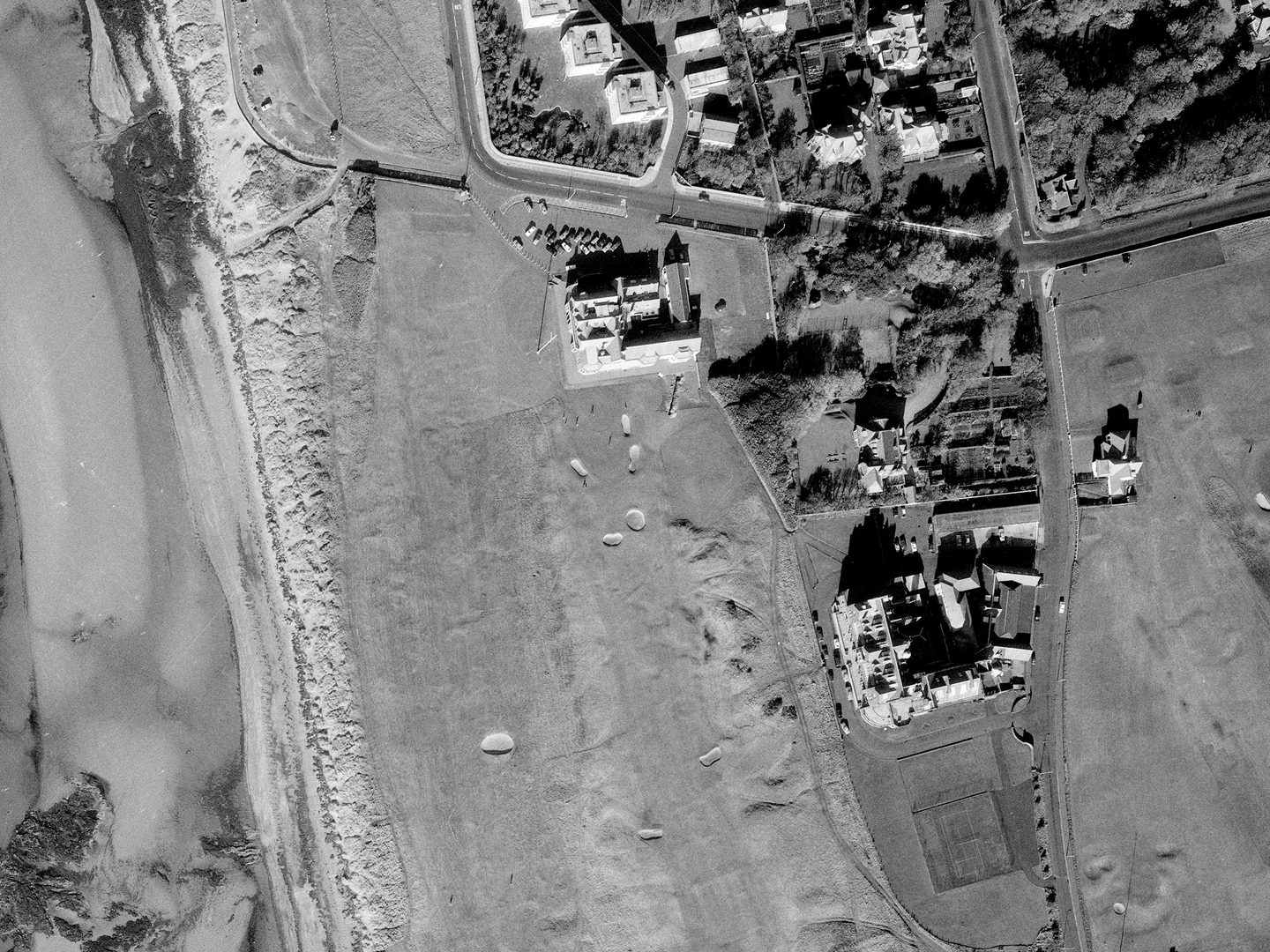 A historic black and white vertical aerial photo of a golf course along a shoreline. The water is to the left of the image, with the course running from top to bottom. Several grand buildings - including the Royal Troon clubhouse - are visible in the centre of the image.