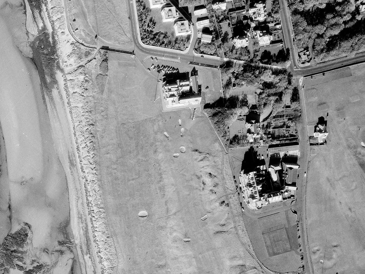 A historic black and white vertical aerial photo of a golf course along a shoreline. The water is to the left of the image, with the course running from top to bottom. Several grand buildings - including the Royal Troon clubhouse - are visible in the centre of the image.