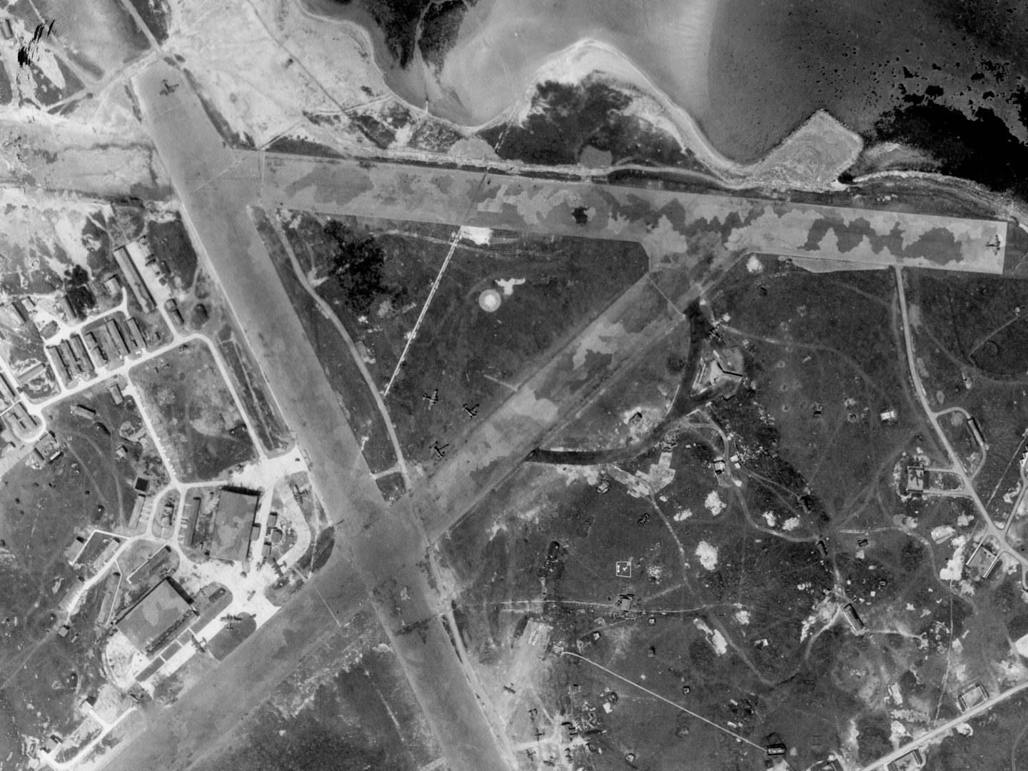 A historic black and white vertical aerial photo of an airfield with 3 runways arranged in a capital A shape. The runways are camouflaged, and several buildings are visible to the left. The coastline is visible towards the top of the image.