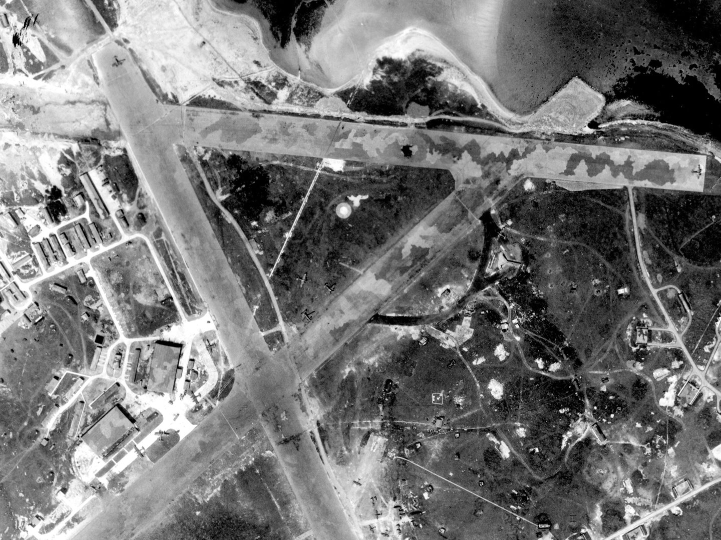 A historic black and white vertical aerial photo of an airfield with 3 runways arranged in a capital A shape. The runways are camouflaged, and several buildings are visible to the left. The coastline is visible towards the top of the image.