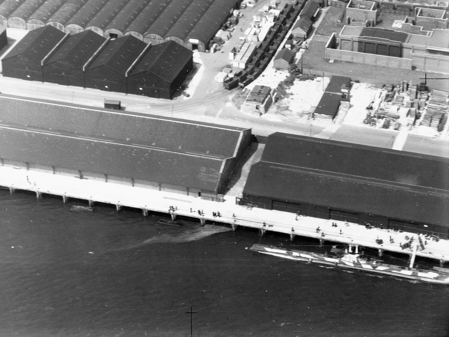 A historic black and white oblique vertical aerial photo of warehouses on a dock. The sea is visible at the front edge of the image. A submarine painted with camouflage colouring is tied up at the dock.