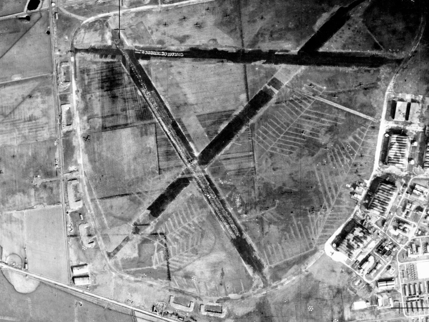 A historic black and white vertical aerial photo of an airfield, with 3 runways arranged in the shape of a capital A. A selection of airfield buildings are visible to the right.
