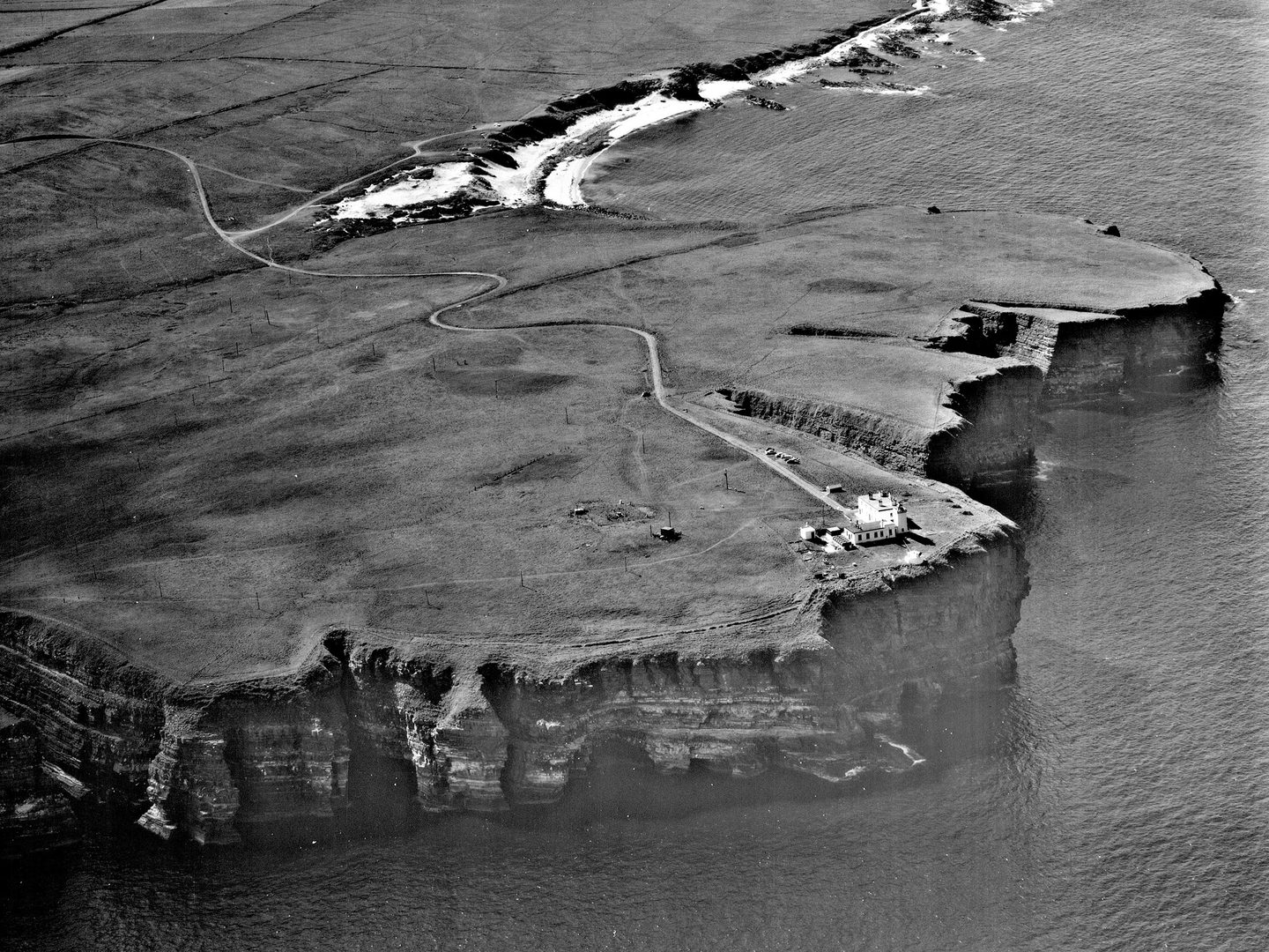 A black and white oblique aerial photo of a headland. A sandy beach stretches into the distance beyond steep cliffs falling into the sea. A white building sits on top of the cliffs. To the building’s left is a square, wire enclosure.