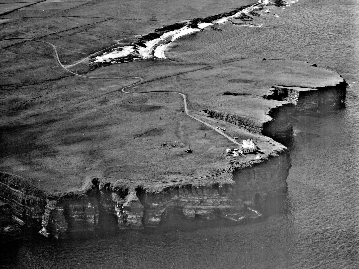 A black and white oblique aerial photo of a headland. A sandy beach stretches into the distance beyond steep cliffs falling into the sea. A white building sits on top of the cliffs. To the building’s left is a square, wire enclosure.