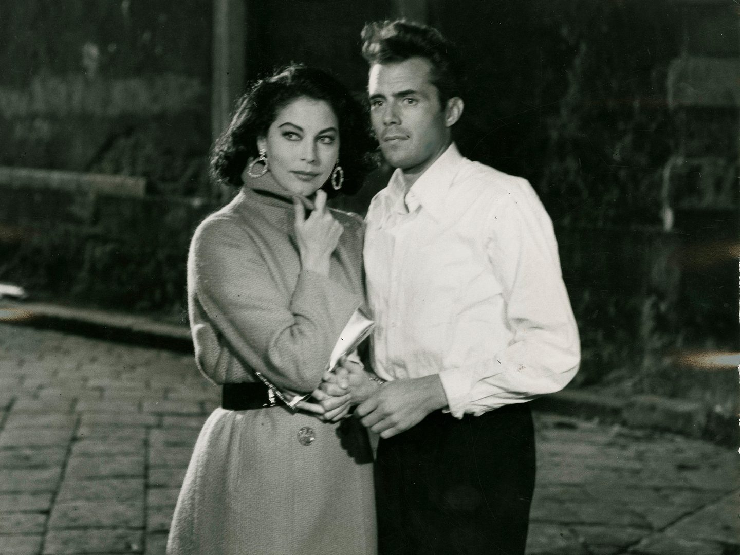 A historic black and white photo of Dirk Bogarde as a young man in a white shirt. Holding his arm to the left is a woman with dark hair in a long coat with her left hand up to her chin.