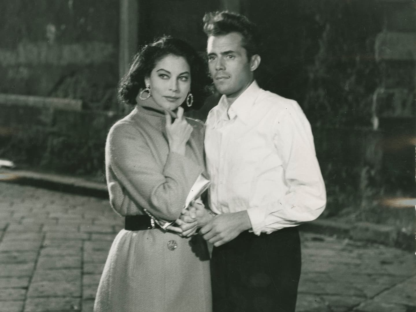 A historic black and white photo of Dirk Bogarde as a young man in a white shirt. Holding his arm to the left is a woman with dark hair in a long coat with her left hand up to her chin.
