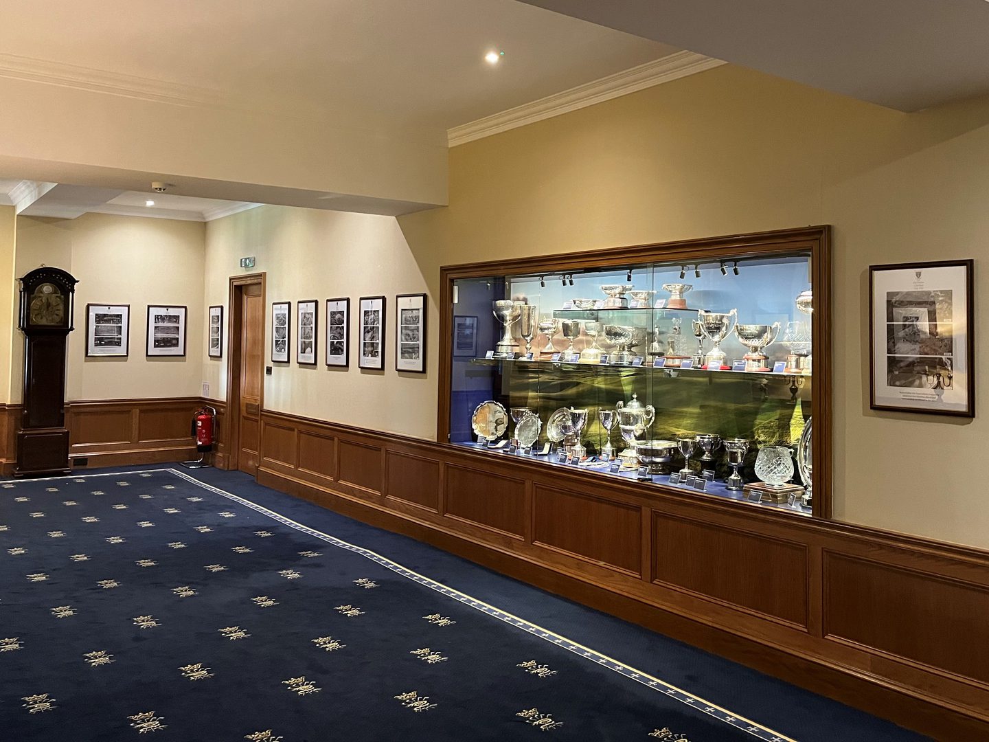 A corridor in a golf clubhouse, with blue carpets and half-panelled walls. A grandfather clock stands to the left and a trophy cabinet is built into the wall to the right. 9 frames showing comparative aerial photos over time line the walls.