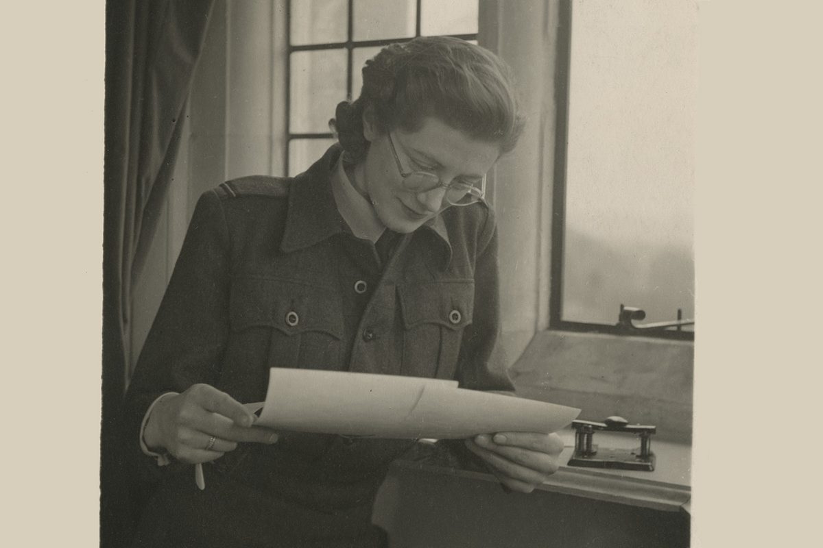 A sepia historic photo of a woman in glasses next to a window. She is looking down at 2 photographic prints in her hands.