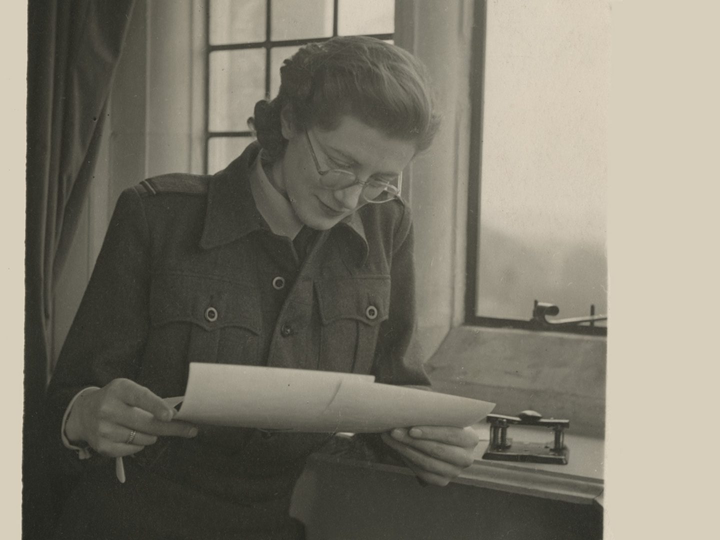 A sepia historic photo of a woman in glasses next to a window. She is looking down at 2 photographic prints in her hands.