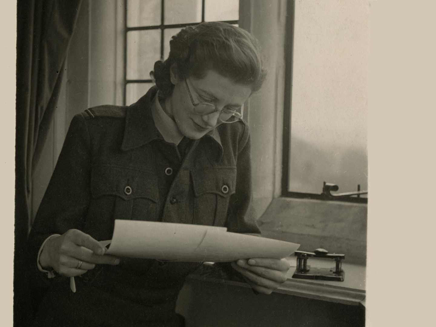 A sepia historic photo of a woman in glasses next to a window. She is looking down at 2 photographic prints in her hands.