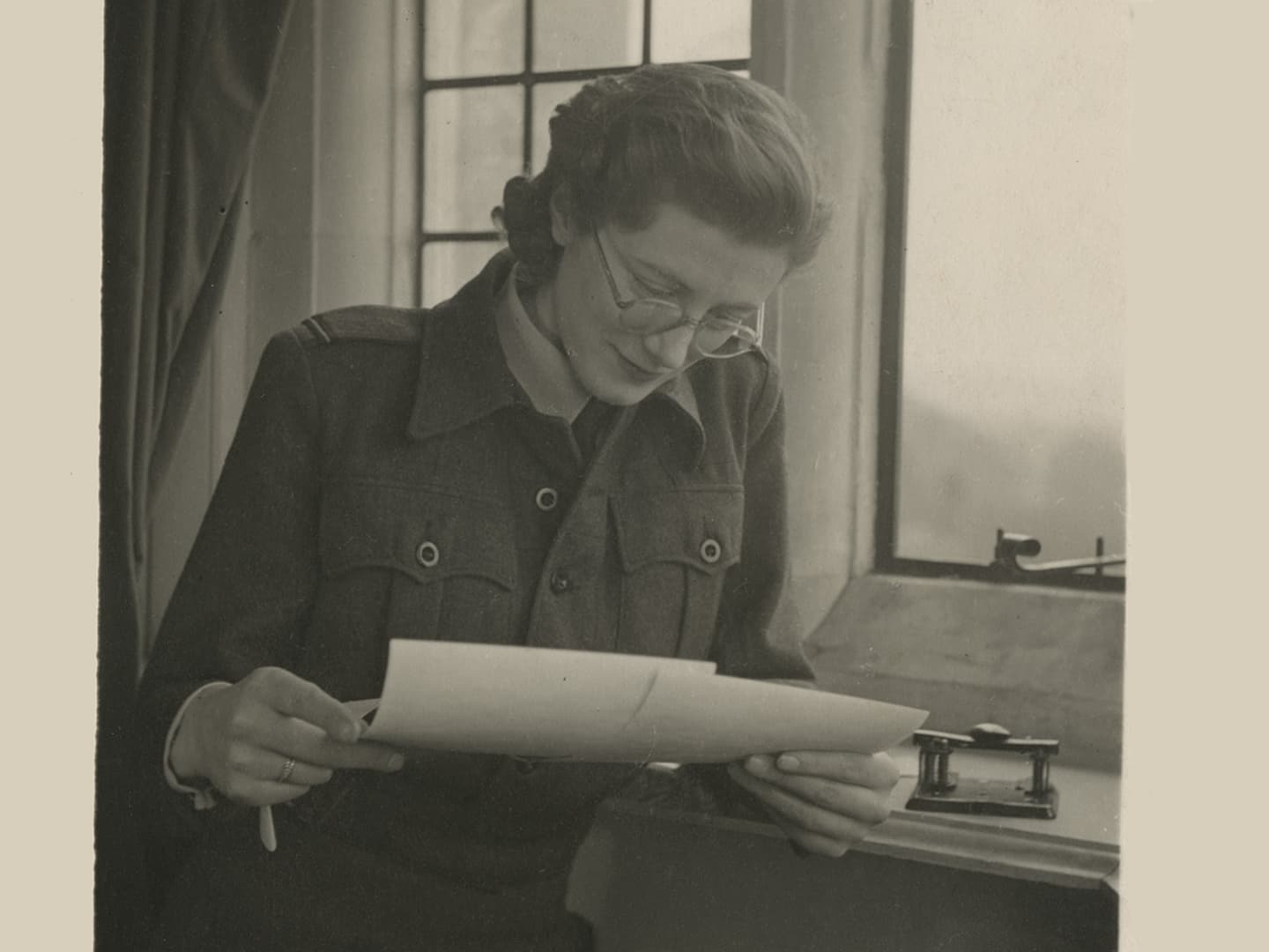 A sepia historic photo of a woman in glasses next to a window. She is looking down at 2 photographic prints in her hands.