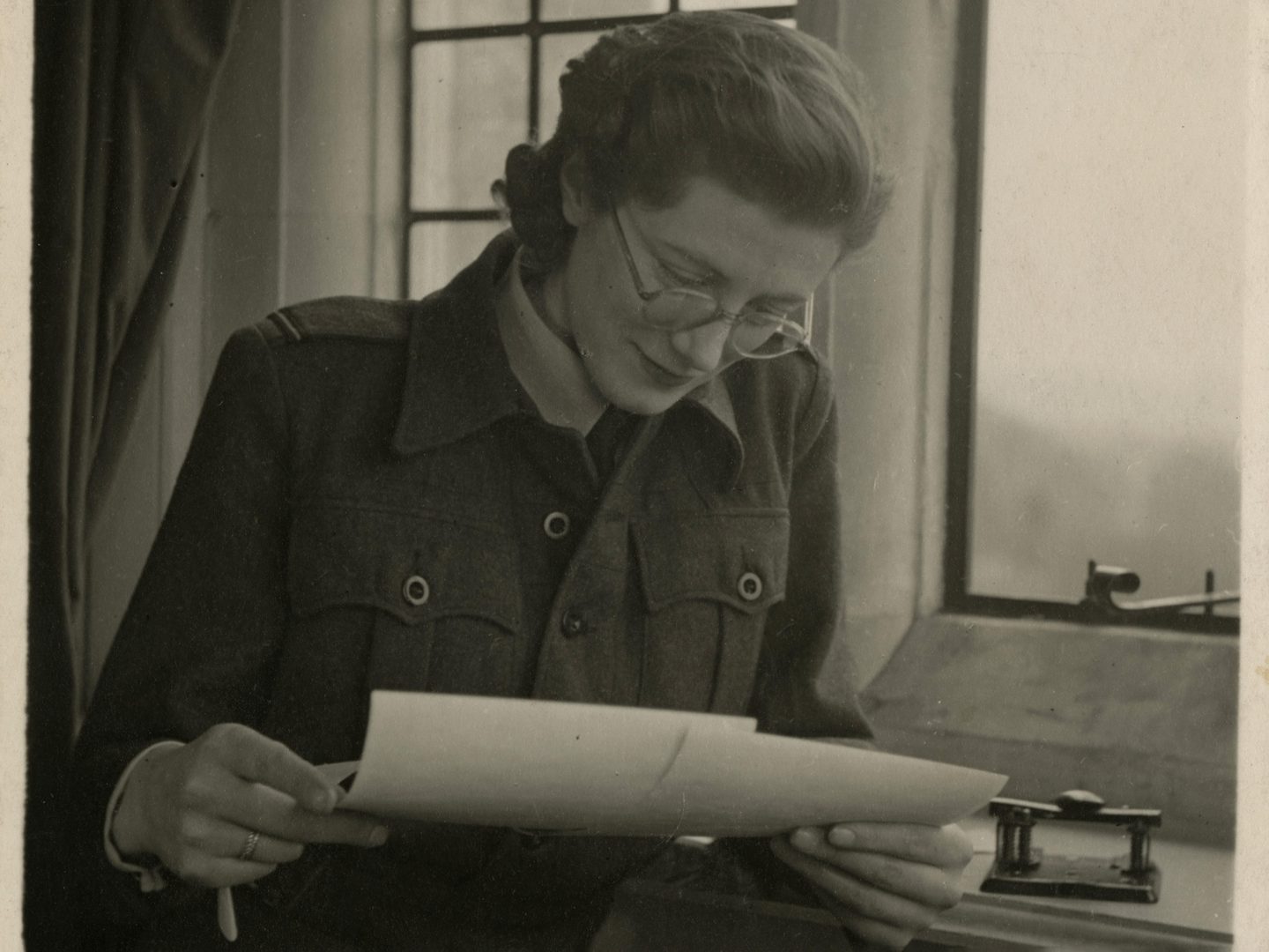 A sepia historic photo of a woman in glasses next to a window. She is looking down at 2 photographic prints in her hands.