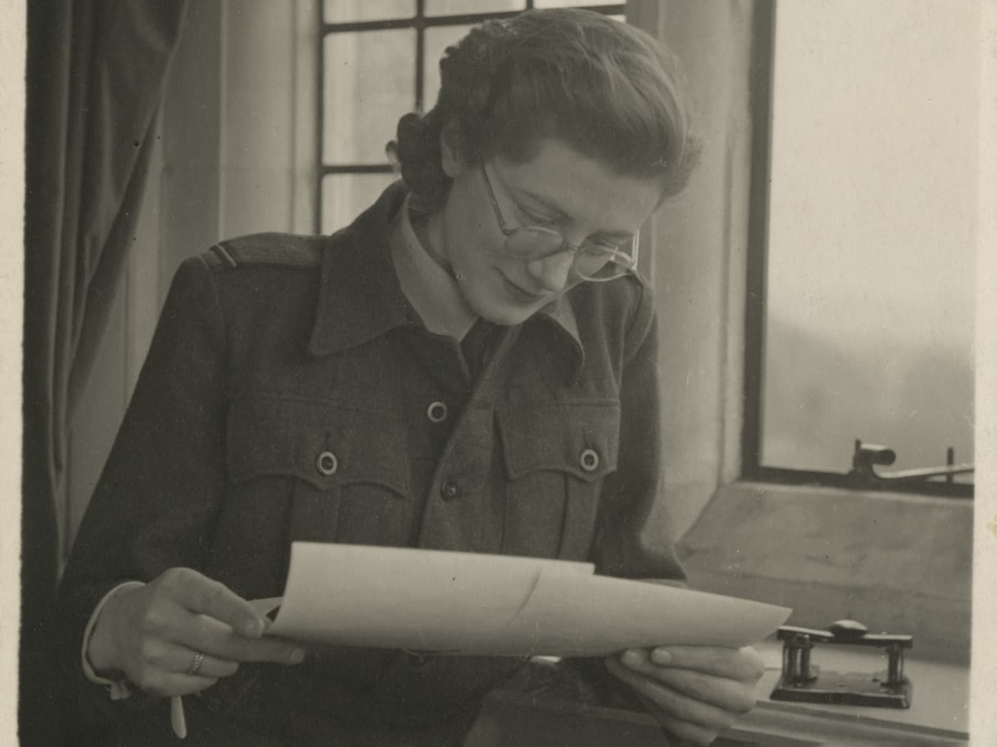 A sepia historic photo of a woman in glasses next to a window. She is looking down at 2 photographic prints in her hands.