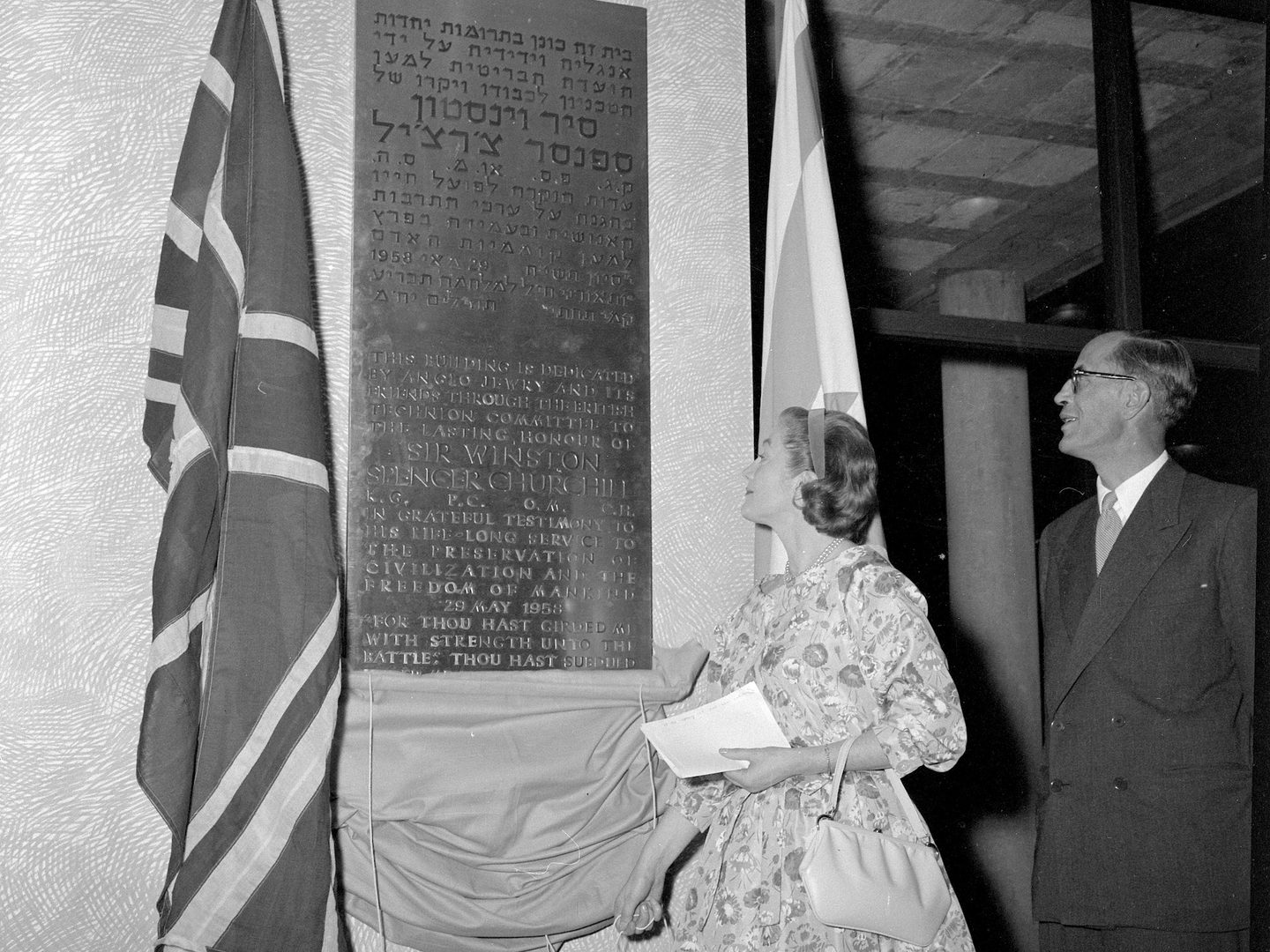 A black and white historic photo of a woman and a man looking up at a plaque. The plaque is engraved with writing in Hebrew and English and flanked by 2 flags.