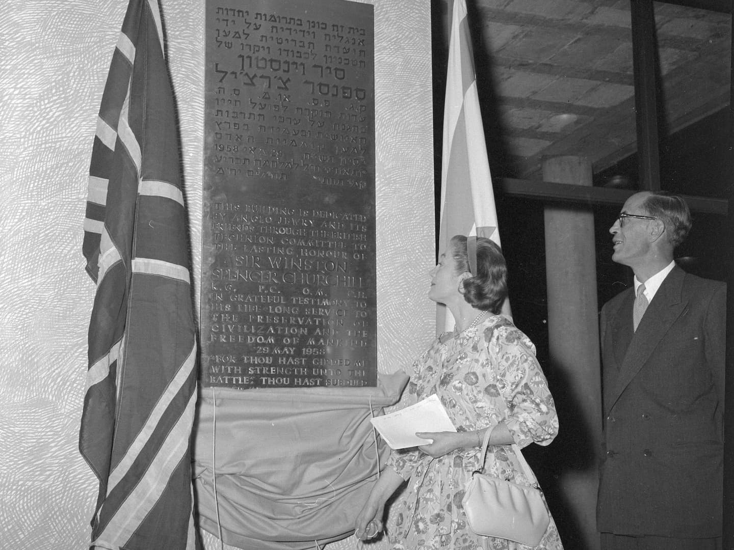 A black and white historic photo of a woman and a man looking up at a plaque. The plaque is engraved with writing in Hebrew and English and flanked by 2 flags.