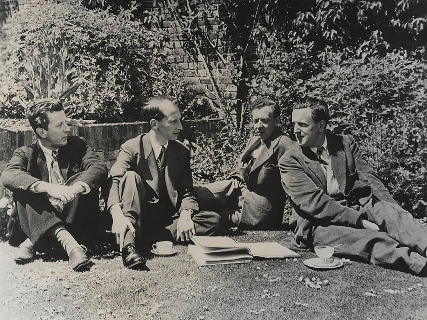 A black and white historic photo of 4 men sitting on a lawn in front of some bushes. They are wearing casual suits. On the grass in front of them are some piles of paperwork, a cup and a glass.