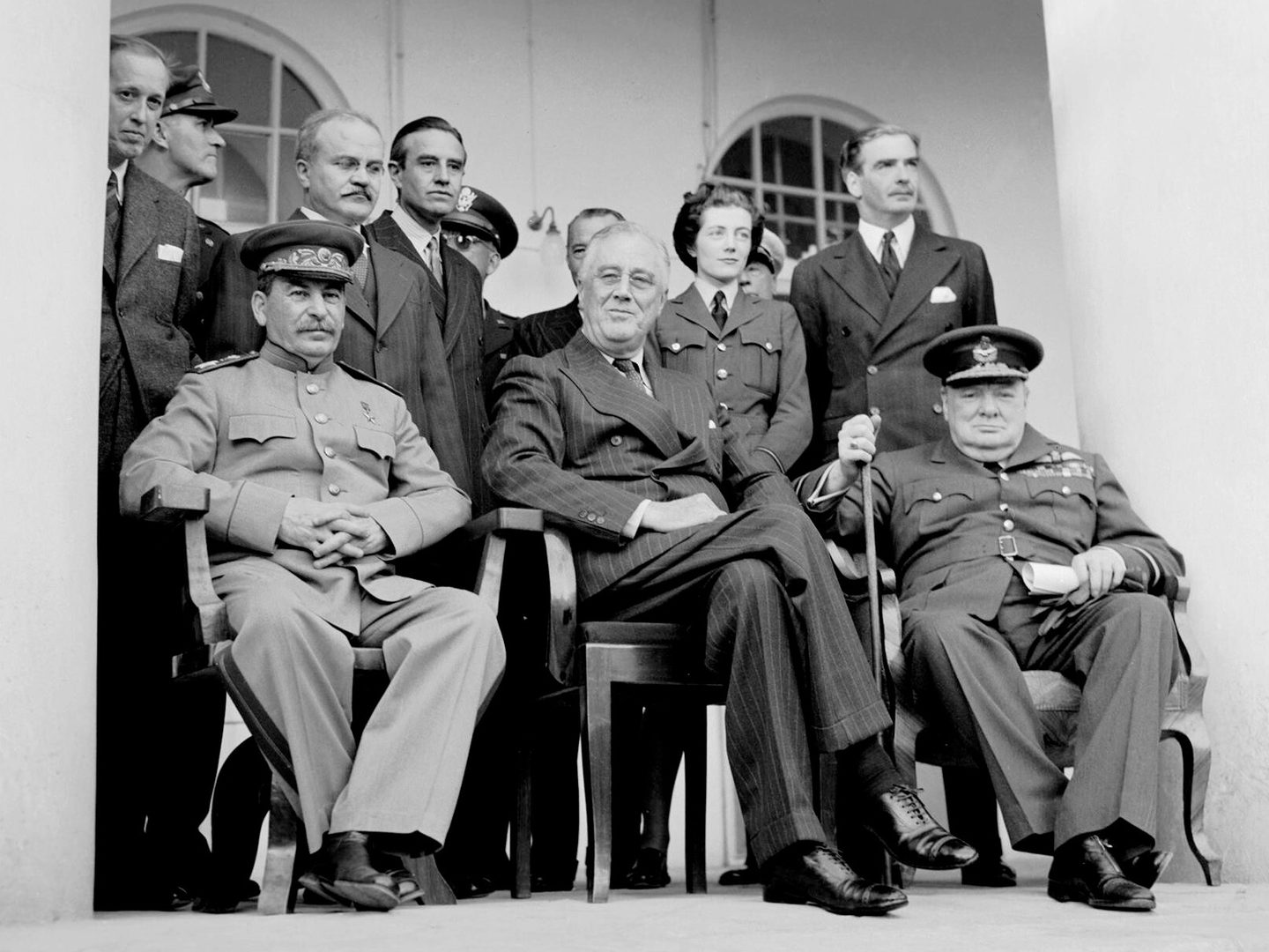A black and white photo of a group of people between 2 white arches. Stalin, Roosevelt and Churchill sit at the front, with Spencer-Churchill standing behind.